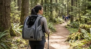 A hiker wearing a gray cat backpack carrier with a curious black cat peering out of the top mesh opening on a forest trail.